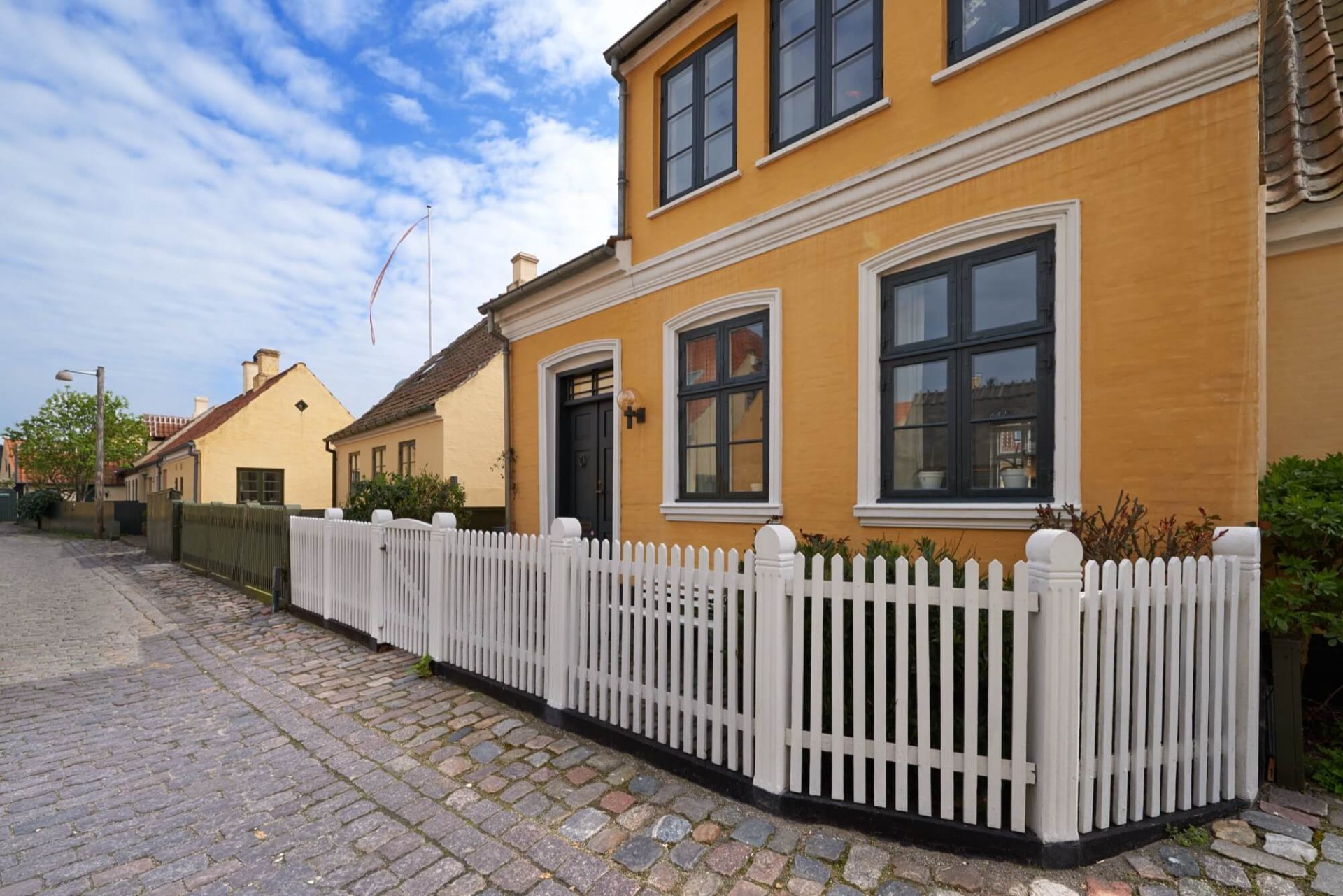 Det står ett anrikt hus med en frappant gul fasad, omgivet av fönster i djupt svart och skyddat av ett vitmålat staket, längs en gata av kullersten